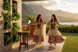 Two hula dancers in grass skirts and floral leis dancing on a Hawaiian terrace at golden hour with Albizia Rose Apple Blossom honey jar on a wooden table, mountains and ocean in the background | HeyHoney.biz