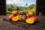 Mango Tropical Fruit Blossom honey jar surrounded by fresh mangoes on a rustic wooden table with a Hawaiian palm tree and mountain landscape in the background | HeyHoney.biz
